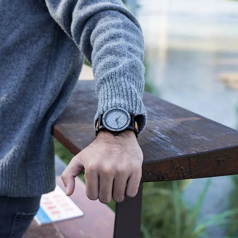 Stylish wood and stone watch on wrist of person wearing gray sweater outdoors near wooden table