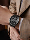 Waterproof luxury watch for men with black dial and metal strap worn with plaid blazer
